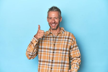 Foto de Hombre caucásico de mediana edad sobre fondo azul levantando ambos pulgares hacia arriba, sonriendo y confiado. - Imagen libre de derechos