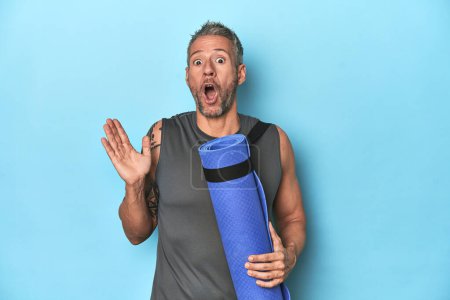Téléchargez les photos : Athlète avec un tapis sur fond de studio bleu surpris et choqué. - en image libre de droit