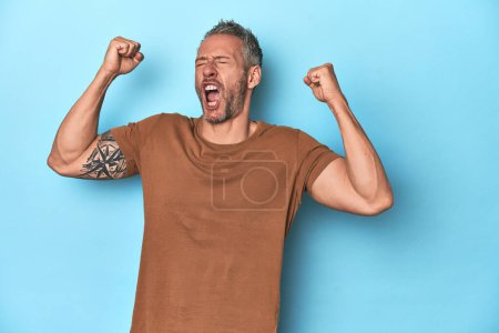 Foto de Hombre caucásico de mediana edad sobre fondo azul levantando puño después de una victoria, concepto ganador. - Imagen libre de derechos