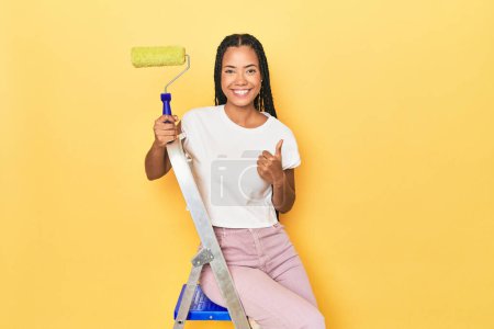 Photo for Indonesian woman with roller on ladder on yellow smiling and raising thumb up - Royalty Free Image