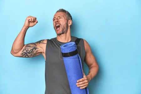 Photo for Athlete with a mat on a blue studio backdrop raising fist after a victory, winner concept. - Royalty Free Image