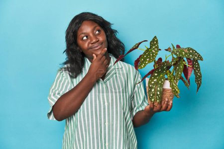 Foto de Curvy gardener holding plant en estudio mirando hacia los lados con expresión dudosa y escéptica. - Imagen libre de derechos
