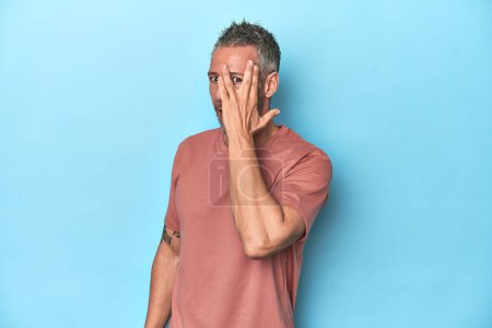 Foto de Hombre caucásico de mediana edad en el telón de fondo azul parpadear en la cámara a través de los dedos, cara cubierta avergonzado. - Imagen libre de derechos