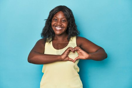 Téléchargez les photos : Jeune femme afro-américaine courbée souriante et montrant une forme de coeur avec les mains. - en image libre de droit
