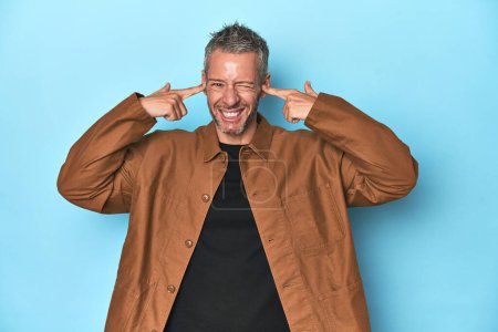 Photo for Middle-aged caucasian man on blue backdrop covering ears with fingers, stressed and desperate by a loudly ambient. - Royalty Free Image