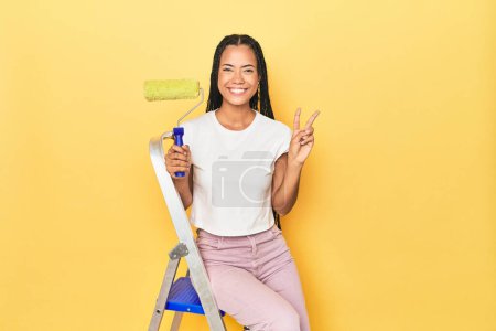 Photo for Indonesian woman with roller on ladder on yellow showing number two with fingers. - Royalty Free Image