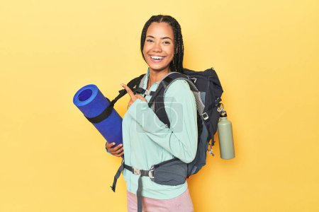 Foto de Mujer indonesia lista para el viaje de montaña en amarillo sonriendo y señalando a un lado, mostrando algo en espacio en blanco. - Imagen libre de derechos