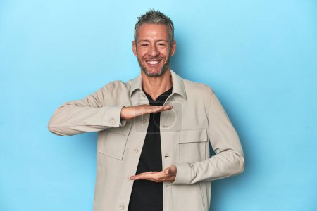 Photo for Middle-aged caucasian man on blue backdrop holding something with both hands, product presentation. - Royalty Free Image