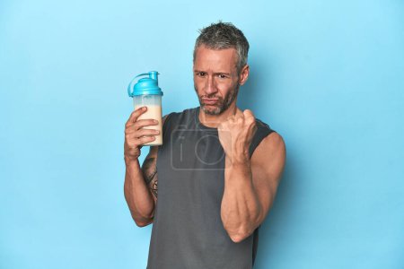 Foto de Atleta sosteniendo batido de proteínas sobre fondo azul mostrando puño a cámara, expresión facial agresiva. - Imagen libre de derechos
