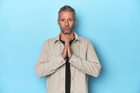 Photo for Middle-aged caucasian man on blue backdrop praying, showing devotion, religious person looking for divine inspiration. - Royalty Free Image