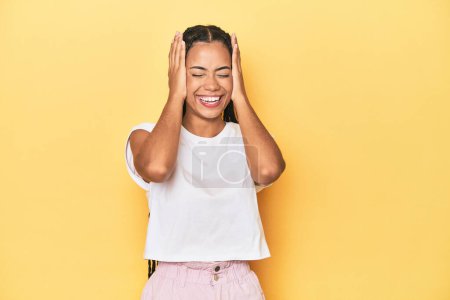 Photo for Young Indonesian woman on yellow studio backdrop laughs joyfully keeping hands on head. Happiness concept. - Royalty Free Image