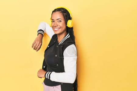 Téléchargez les photos : Femme indonésienne en veste de baseball avec écouteurs sur fond de studio jaune - en image libre de droit