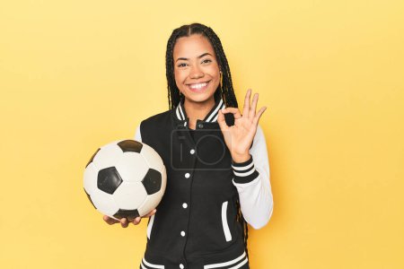 Photo for Indonesian schoolgirl with soccer ball on yellow cheerful and confident showing ok gesture. - Royalty Free Image