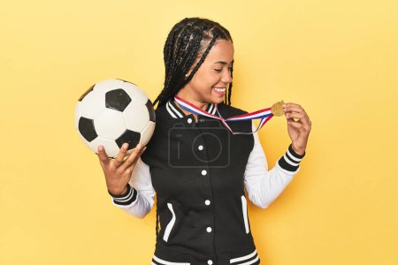 Téléchargez les photos : Jeune Indonésienne avec médaille et ballon de football sur fond de studio jaune - en image libre de droit