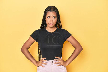 Photo for Young Indonesian woman on yellow studio backdrop confused, feels doubtful and unsure. - Royalty Free Image