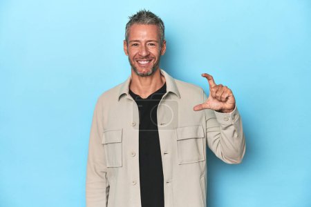 Photo for Middle-aged caucasian man on blue backdrop holding something little with forefingers, smiling and confident. - Royalty Free Image