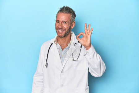 Photo for Caucasian middle-aged doctor on blue background cheerful and confident showing ok gesture. - Royalty Free Image