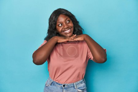 Photo for Young african american curvy woman keeps hands under chin, is looking happily aside. - Royalty Free Image