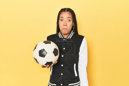 Photo for Indonesian schoolgirl with soccer ball on yellow shrugs shoulders and open eyes confused. - Royalty Free Image