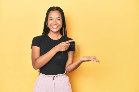 Téléchargez les photos : Jeune femme indonésienne sur fond jaune studio excité tenant un espace de copie sur la paume. - en image libre de droit
