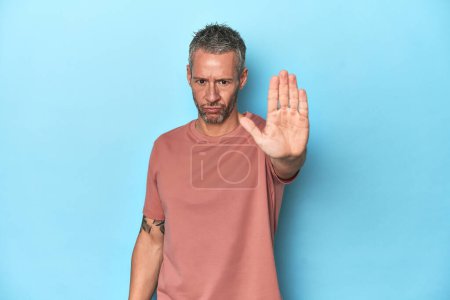 Photo for Middle-aged caucasian man on blue backdrop standing with outstretched hand showing stop sign, preventing you. - Royalty Free Image