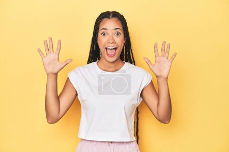 Foto de Joven mujer indonesia en el fondo del estudio amarillo recibiendo una agradable sorpresa, emocionada y levantando las manos. - Imagen libre de derechos
