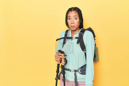 Photo for Indonesian woman with hiking gear on yellow shrugs shoulders and open eyes confused. - Royalty Free Image