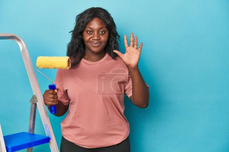 Téléchargez les photos : Femme courbée avec rouleau de peinture et échelle souriant gai montrant numéro cinq avec les doigts. - en image libre de droit
