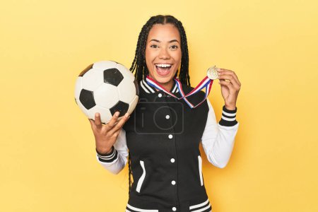 Foto de Joven mujer indonesia con medalla y pelota de fútbol sobre fondo amarillo estudio - Imagen libre de derechos