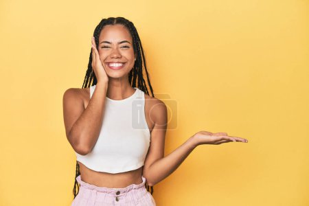 Photo for Young Indonesian woman on yellow studio backdrop holds copy space on a palm, keep hand over cheek. Amazed and delighted. - Royalty Free Image
