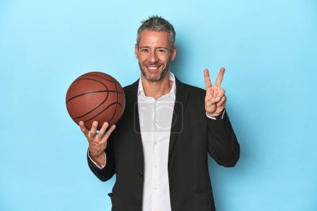 Foto de Entrenador de baloncesto de mediana edad con telón de fondo azul Entrenador de baloncesto de mediana edad con telón de fondo azul que muestra el número dos con los dedos. - Imagen libre de derechos
