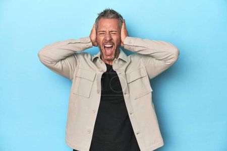Photo for Middle-aged caucasian man on blue backdrop covering ears with hands trying not to hear too loud sound. - Royalty Free Image