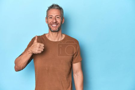 Foto de Hombre caucásico de mediana edad sobre fondo azul sonriendo y levantando el pulgar hacia arriba - Imagen libre de derechos