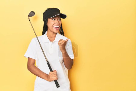 Photo for Indonesian female golfer on yellow backdrop points with thumb finger away, laughing and carefree. - Royalty Free Image