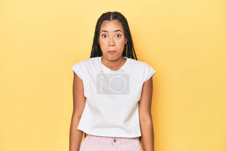 Photo for Young Indonesian woman on yellow studio backdrop shrugs shoulders and open eyes confused. - Royalty Free Image