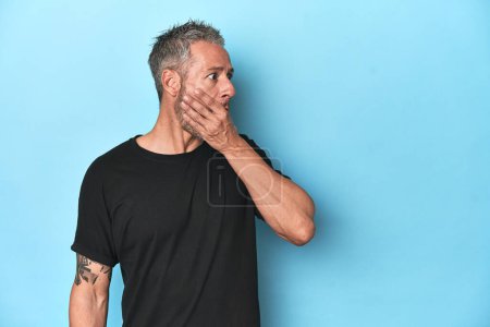 Foto de Hombre caucásico de mediana edad sobre fondo azul reflexivo mirando a un espacio de copia que cubre la boca con la mano. - Imagen libre de derechos