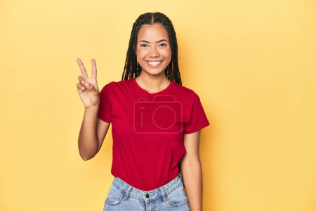Photo for Young Indonesian woman on yellow studio backdrop joyful and carefree showing a peace symbol with fingers. - Royalty Free Image