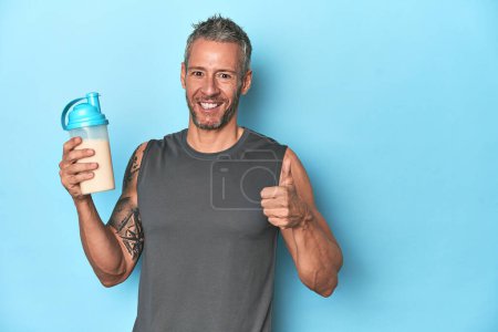 Photo for Athlete holding protein shake on blue backdrop smiling and raising thumb up - Royalty Free Image