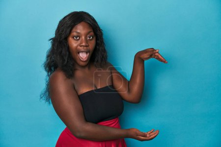 Foto de Joven afroamericana curvilínea mujer sorprendida y asombrada sosteniendo un espacio de copia entre manos. - Imagen libre de derechos