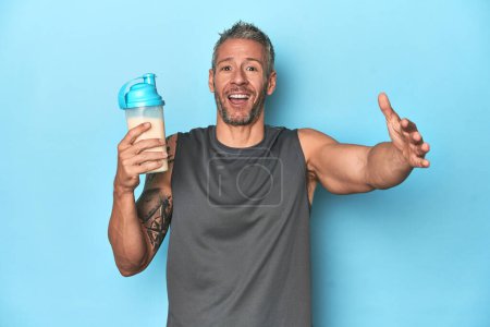Photo for Athlete holding protein shake on blue backdrop receiving a pleasant surprise, excited and raising hands. - Royalty Free Image