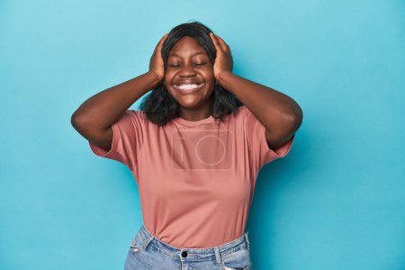 Foto de Joven mujer con curvas afroamericana se ríe alegremente manteniendo las manos en la cabeza. Concepto de felicidad. - Imagen libre de derechos