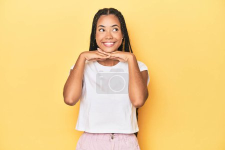 Photo for Young Indonesian woman on yellow studio backdrop keeps hands under chin, is looking happily aside. - Royalty Free Image