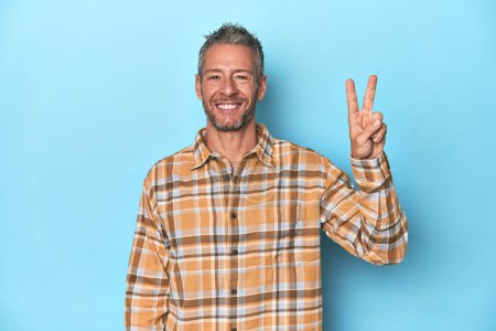Foto de Hombre caucásico de mediana edad sobre fondo azul alegre y despreocupado mostrando un símbolo de paz con los dedos. - Imagen libre de derechos