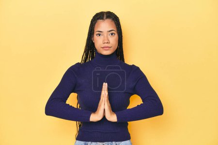 Foto de Joven mujer indonesia en el fondo del estudio amarillo rezando, mostrando devoción, persona religiosa buscando inspiración divina. - Imagen libre de derechos