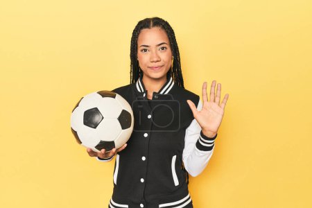Téléchargez les photos : Écolière indonésienne avec ballon de football sur jaune souriant gai montrant numéro cinq avec les doigts. - en image libre de droit
