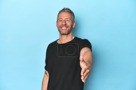 Photo for Middle-aged caucasian man on blue backdrop stretching hand at camera in greeting gesture. - Royalty Free Image