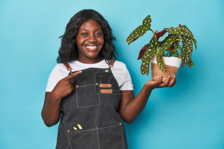 Foto de Curvy jardinero sosteniendo planta en el estudio sonriendo y señalando a un lado, mostrando algo en el espacio en blanco. - Imagen libre de derechos