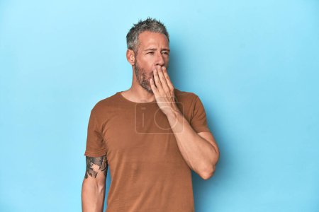 Foto de Hombre caucásico de mediana edad sobre fondo azul bostezando mostrando un gesto cansado cubriendo la boca con la mano. - Imagen libre de derechos