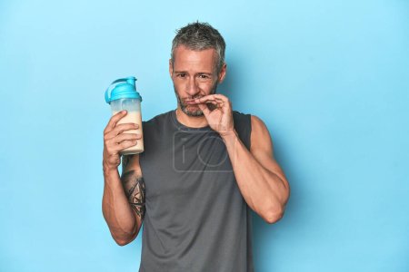 Foto de Atleta sosteniendo batido de proteínas en el fondo azul con los dedos en los labios manteniendo un secreto. - Imagen libre de derechos