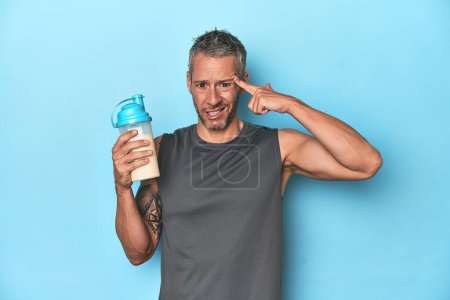 Photo for Athlete holding protein shake on blue backdrop showing a disappointment gesture with forefinger. - Royalty Free Image